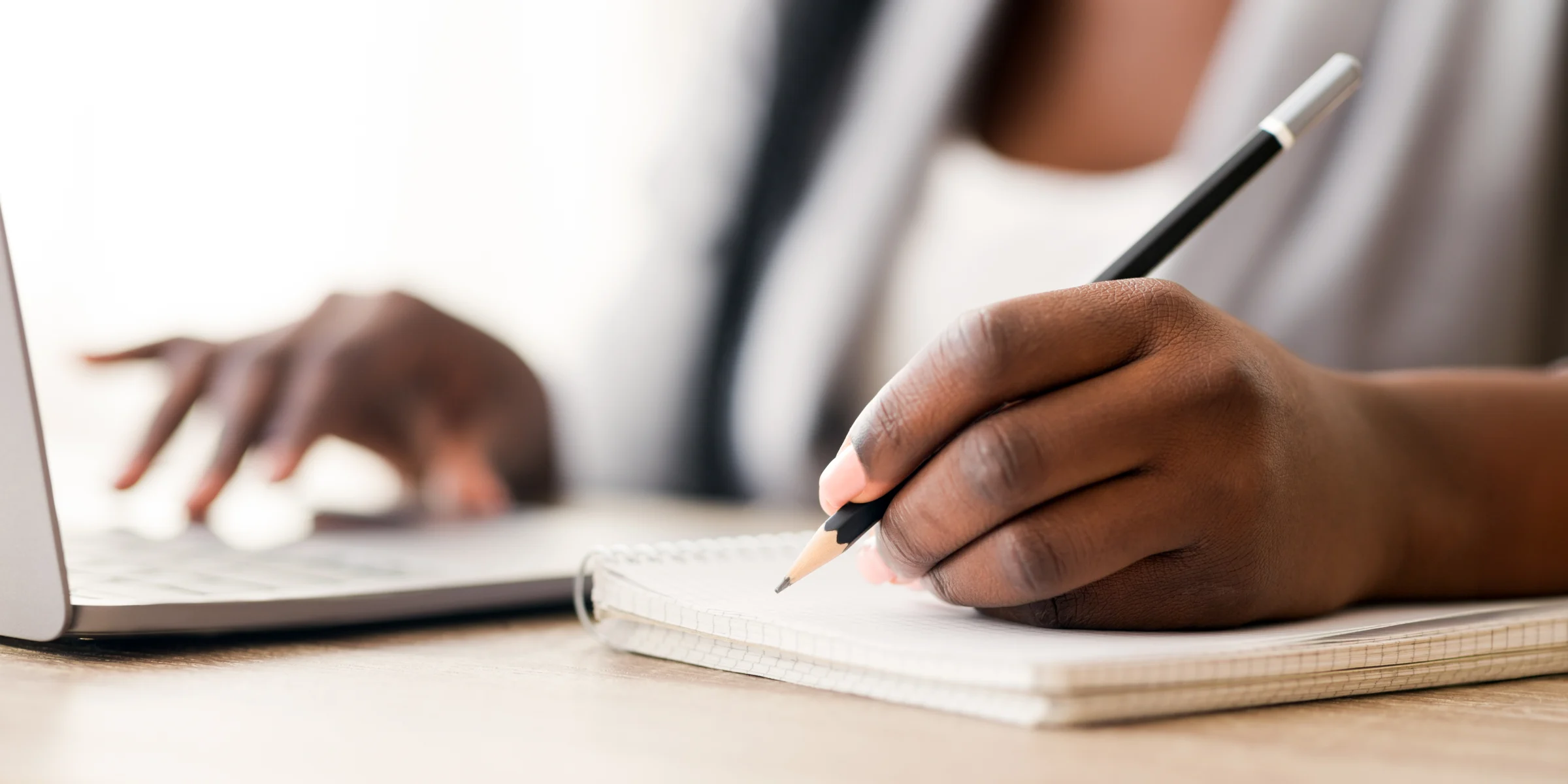 Hands writing in a notebook beside an open laptop